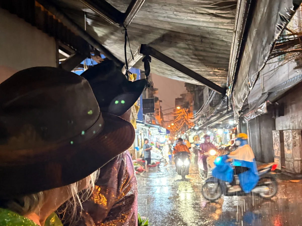 A busy District 4 neighborhood lane during heavy evening rain with motorbikes and local shops