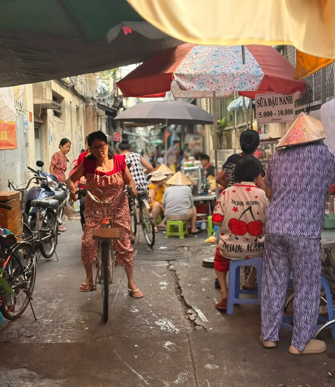 A woman with a bicycle among neighbors and food stalls in a lively District 4 alley, Saigon