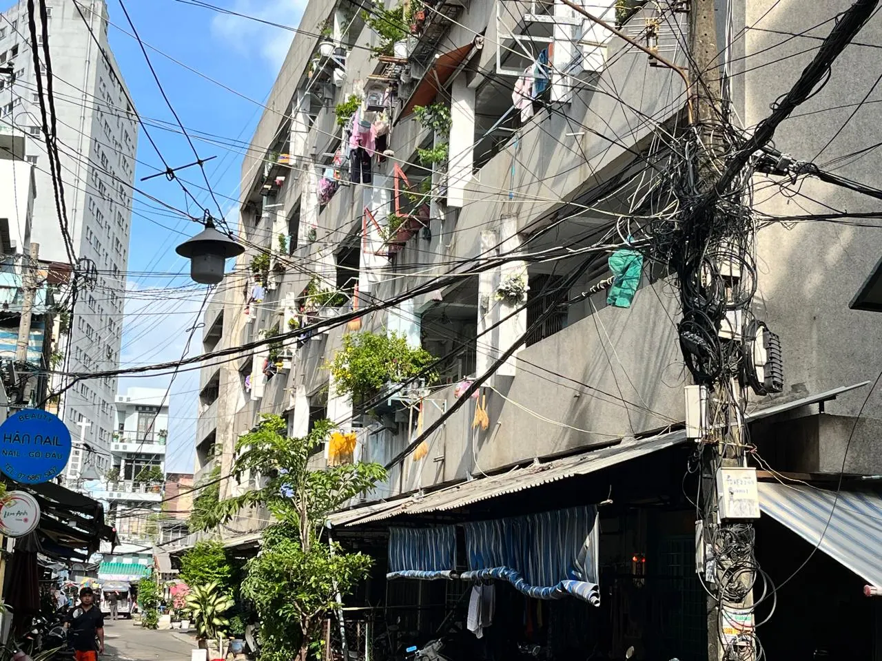 A utility pole crowded with cables beside older apartment buildings in a sunlit District 4 alley, Saigon